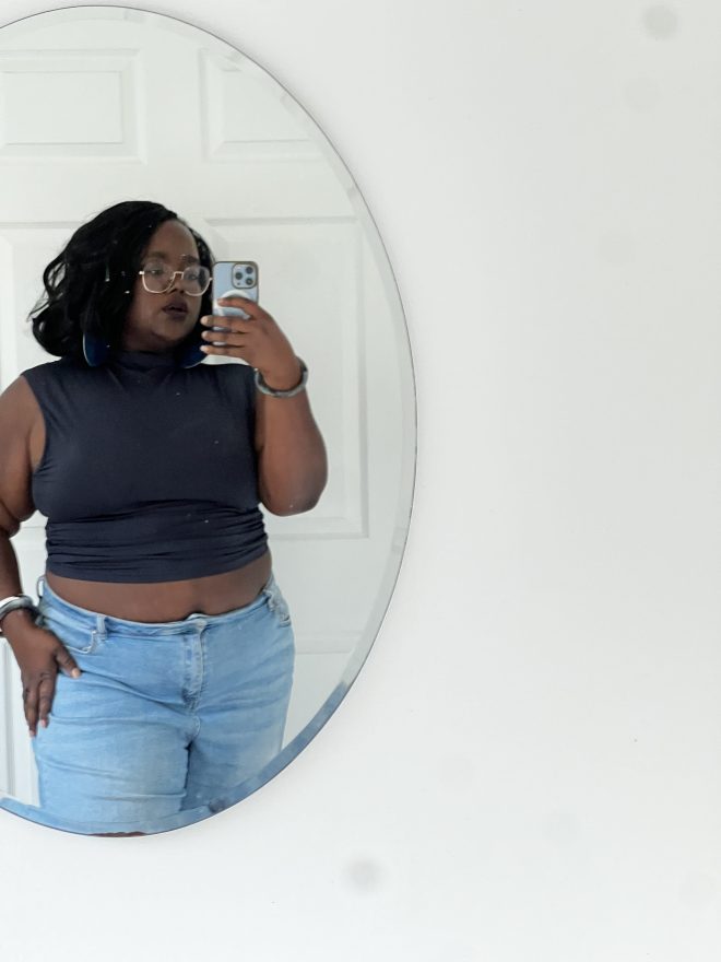 dark skin black woman with a bob hair cut, wearing dark lipstick, navy blue mock neck top and light blue jeans taking the picture in the mirror with her phone.
